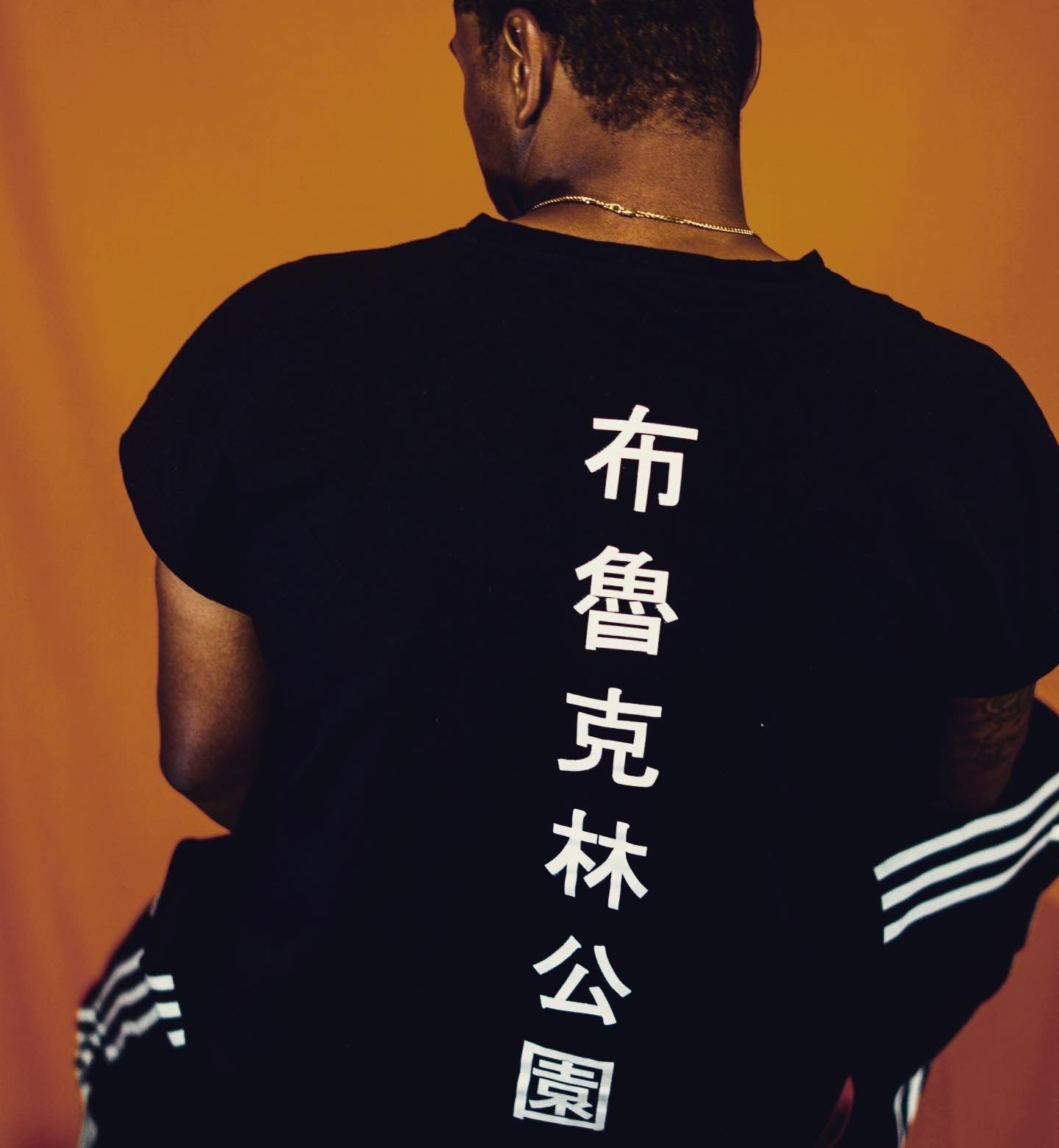 Person facing away from the camera wearing a black T-shirt with white vertical Chinese characters down the back, standing against an orange background.