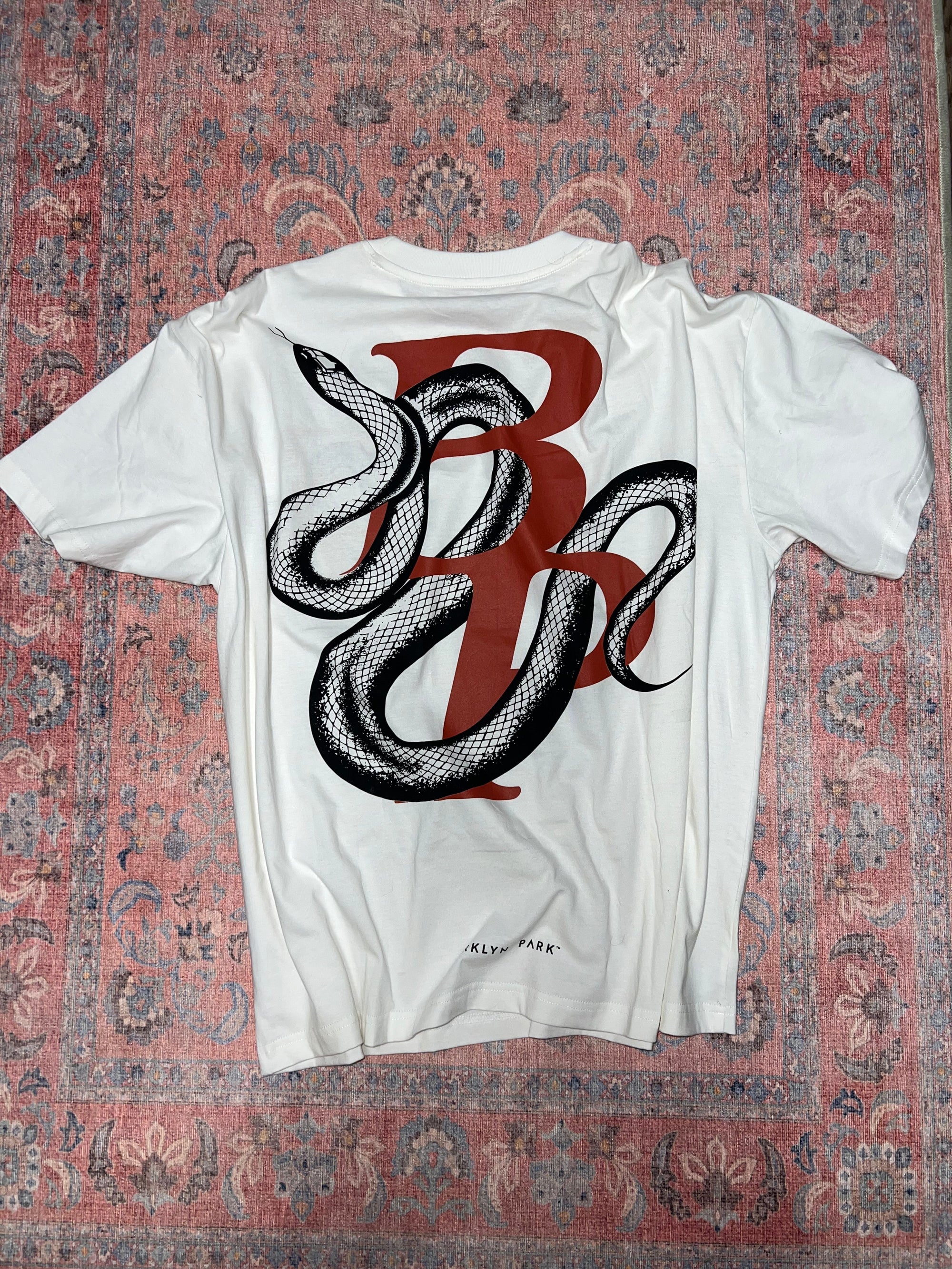 Flat lay of Brklyn Park’s black Snake Tee featuring a bold snake graphic print, displayed on a Bridgerton rug. Part of the Fall 2025 drop, highlighting quiet luxury and elevated streetwear.