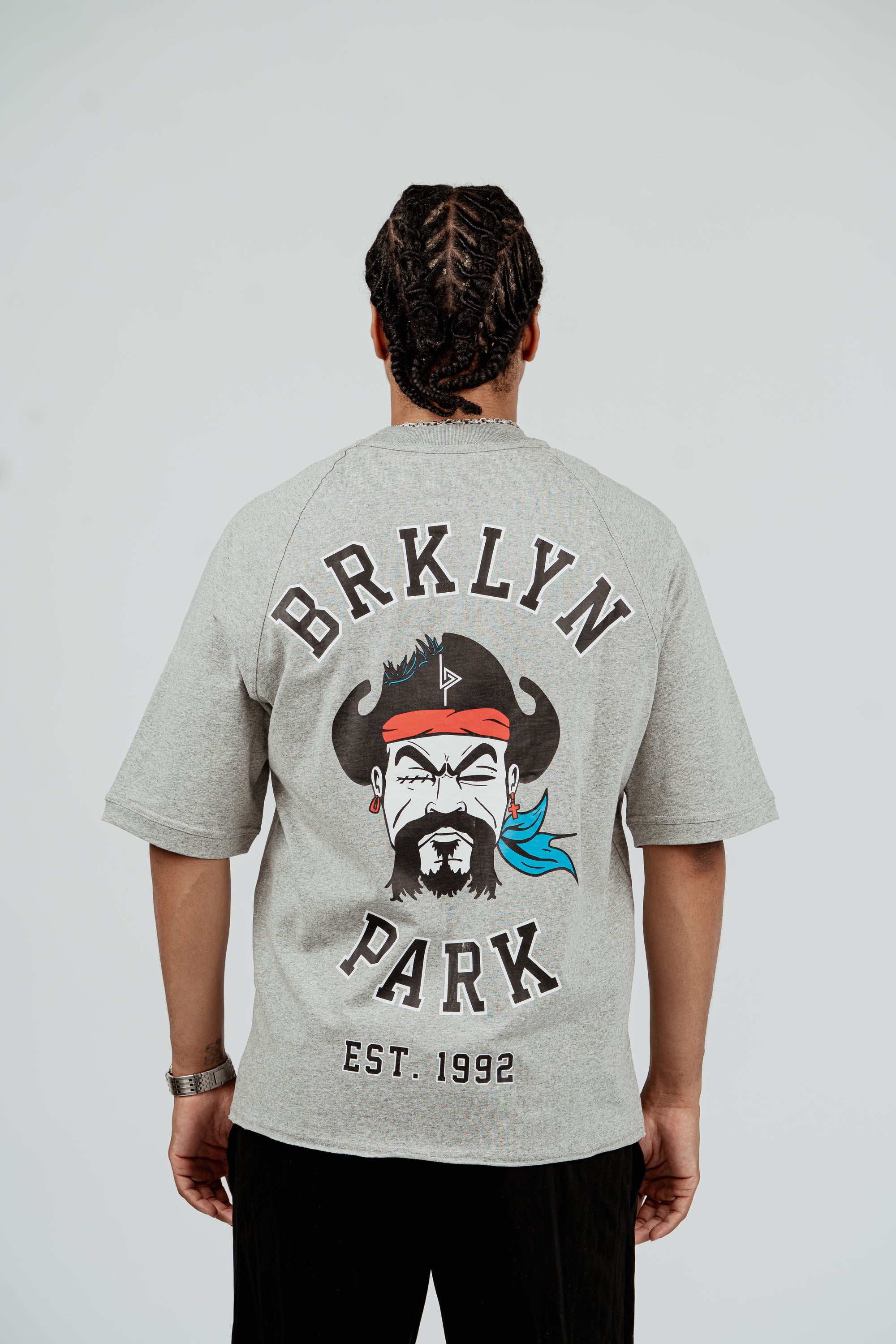 Back view of model wearing Brklyn Park Sanchez Tee in gray featuring large graphic with Brklyn Park logo and illustrated character