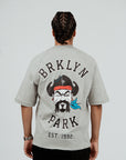 Back view of model wearing Brklyn Park Sanchez Tee in gray featuring large graphic with Brklyn Park logo and illustrated character