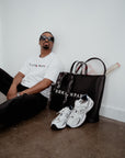 Man sitting on the floor with white sneakers, a black bag, and a tennis racket against a white wall.
