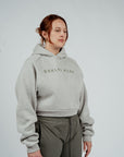 A female model wearing a gray cropped hoodie with 'BRKLYN PARK' logo print on the front, paired with dark pants minimalist streetwear top women’s cropped hoodie neutral loungewear hoodie Brklyn Park hoodie beige cropped hoodie