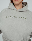 A female model wearing a gray cropped hoodie with 'BRKLYN PARK' logo print on the front, paired with dark pants minimalist streetwear top women’s cropped hoodie neutral loungewear hoodie Brklyn Park hoodie beige cropped hoodie
