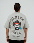 Back view of model wearing Brklyn Park Sanchez Tee in gray featuring large graphic with Brklyn Park logo and illustrated character